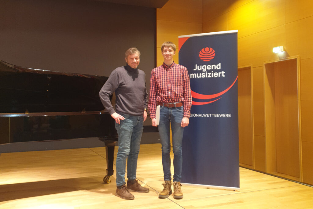 Julian Q. Schüler der Musikschule und Peter Neuhof beim Jugend Musiziert Wettbewerb 2023 stehend vor Banner des Wettbewerbs